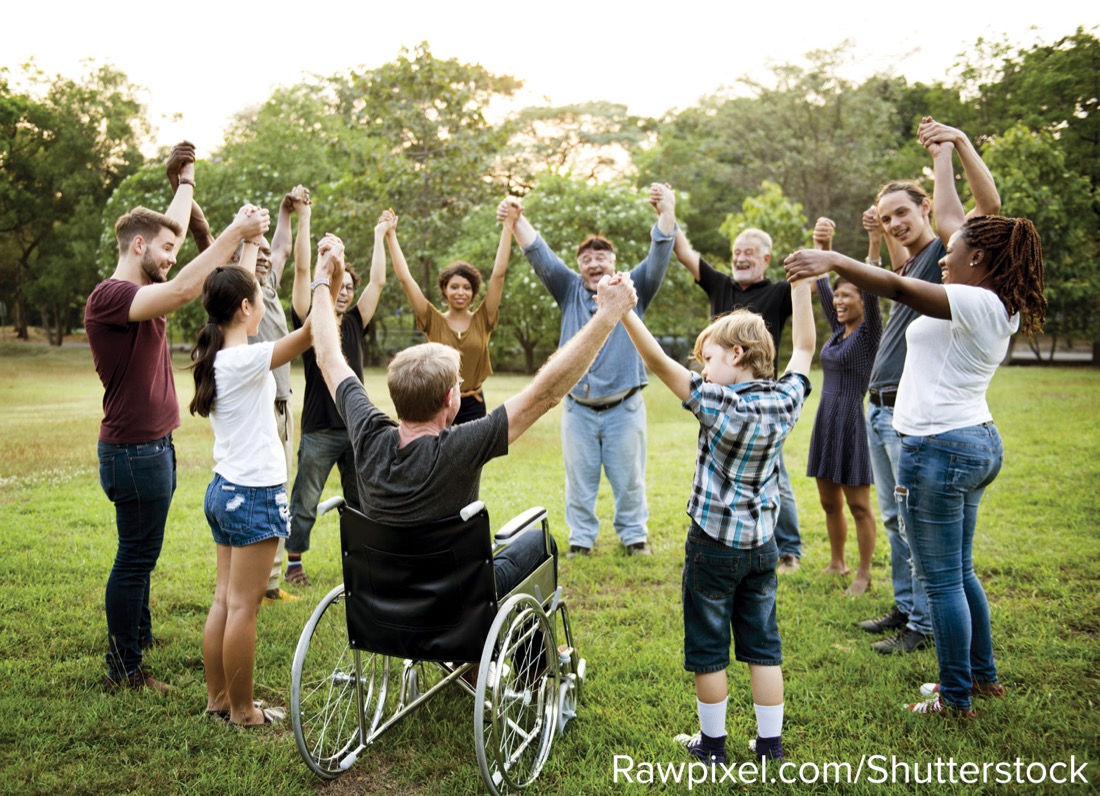 A group of people of different ages standing in a circle outdoors with their hands raised together, one using a wheelchair.