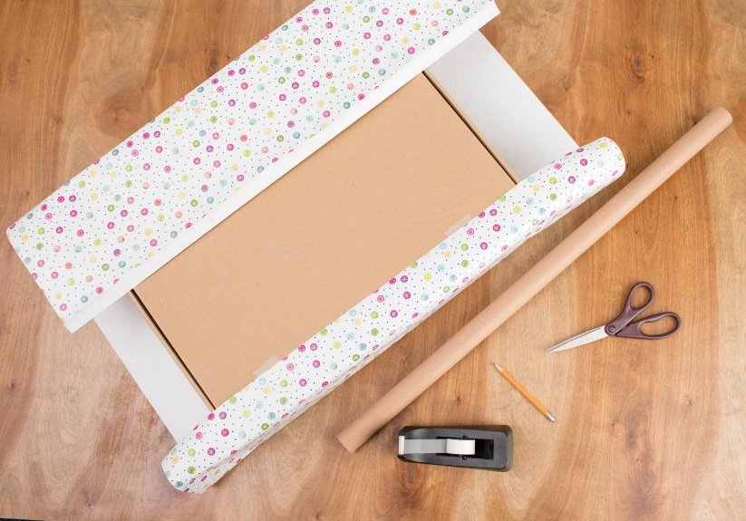 A photograph of a roll of wrapping paper, scissors, a pencil and a tape measure on a wooden floor.